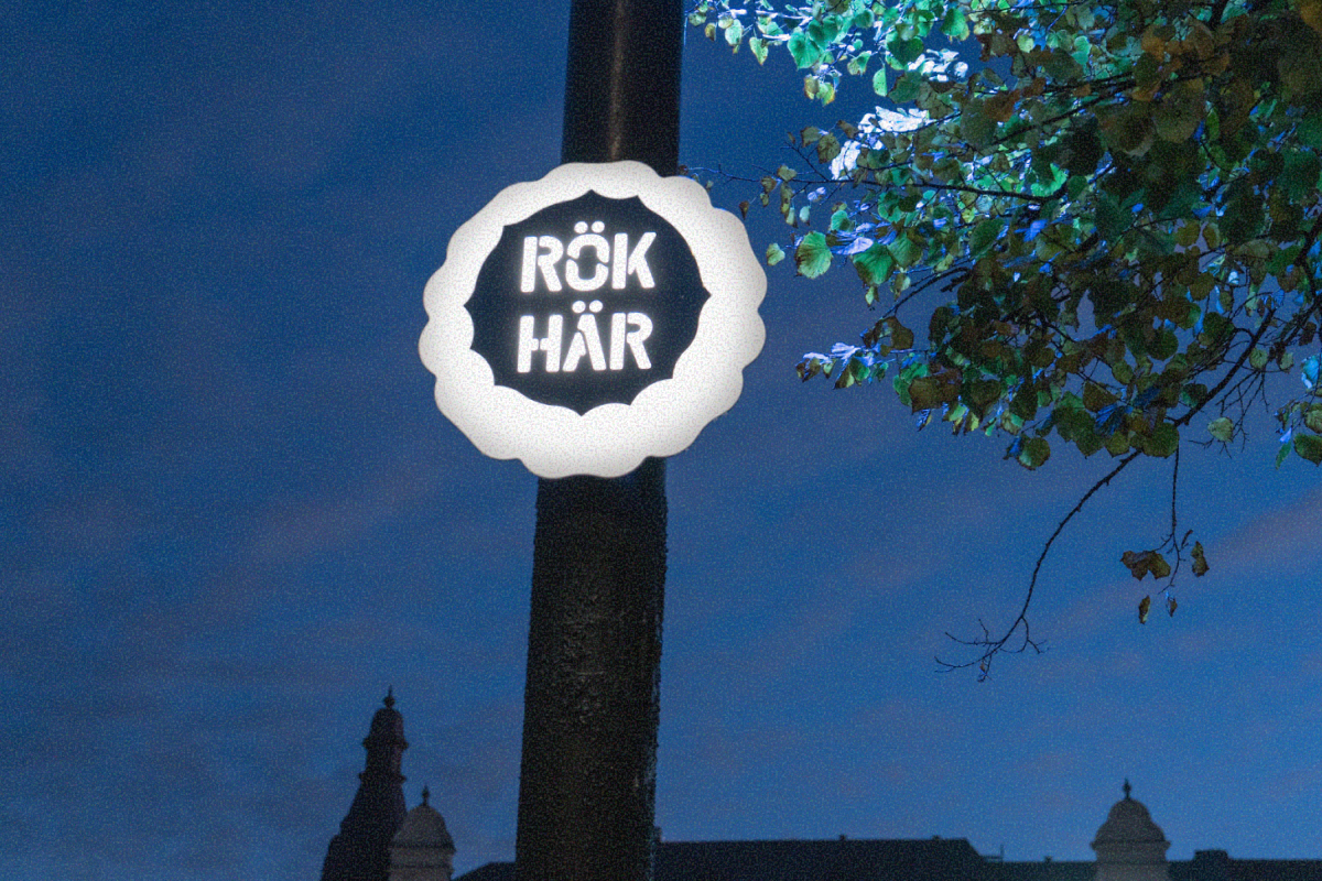 Illuminated round sign with text 'RÖK HÄR' mounted on a pole at dusk with tree branches and building silhouettes in the background.