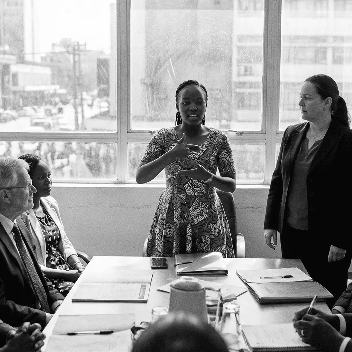 A deaf woman signing with her interpreter next to her, standing up and presenting in a work meeting.