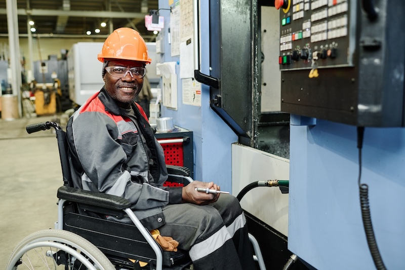 Smiling factory worker in safety gear seated in a wheelchair operating machinery in an industrial setting.