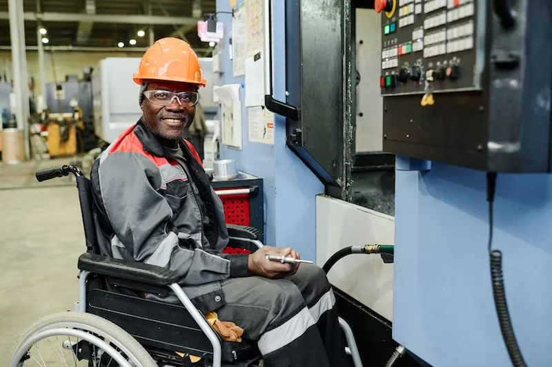 Smiling factory worker in safety gear seated in a wheelchair operating machinery in an industrial setting.