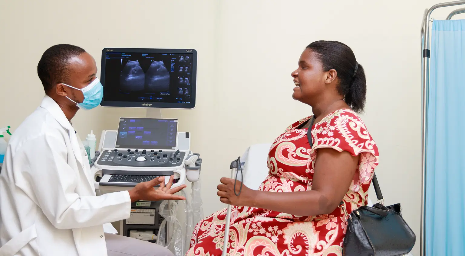 A blind woman is pregnant at a doctor appointment. The doctor is wearing a mouth mask, and he is in front of a computer and talking to the woman.