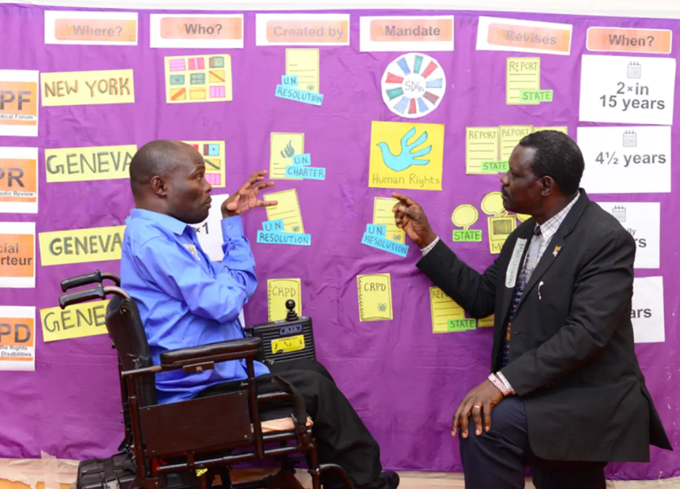 There are two men speaking. One of them is a wheelchair user; the second one is a person with psychosocial disabilities, and he is pointing to some drawings in a panel.