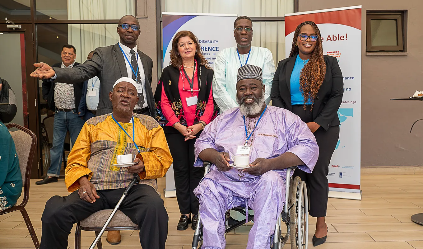 Group of six diverse adults posing at an event with two men seated, one in wheelchair, holding cups.
