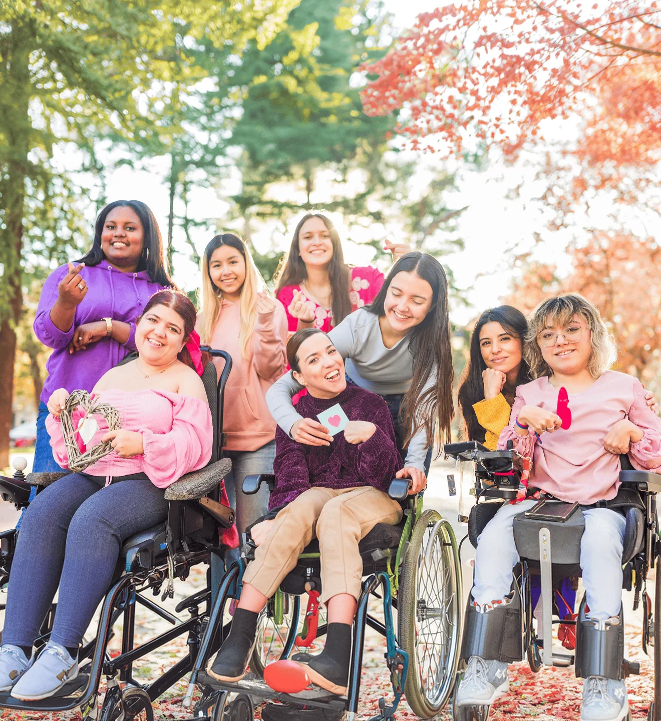 Group of smiling women, including three in wheelchairs, outdoors holding heart symbols.