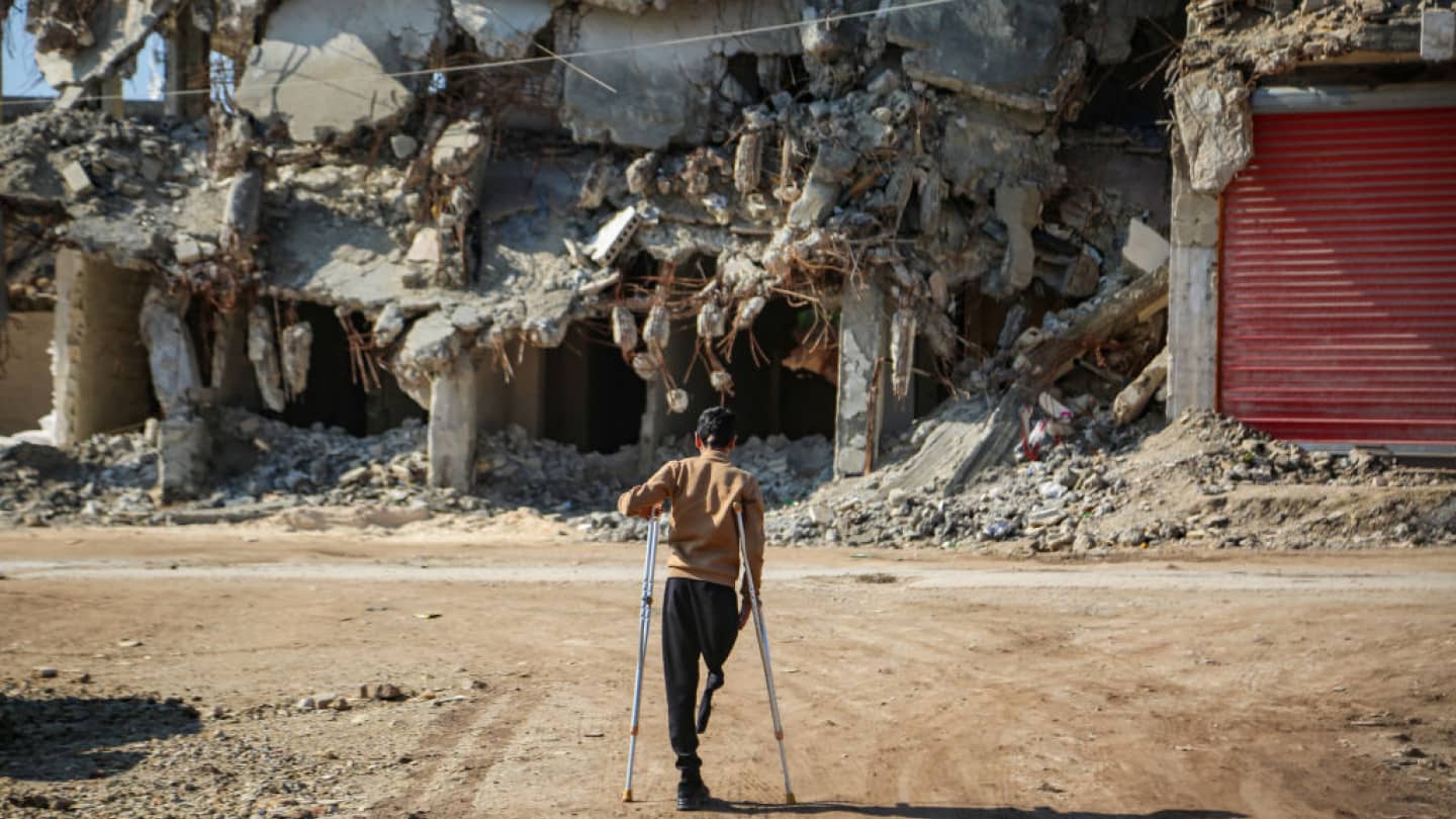 A young amputee boy with a physical disability is using crutches is walking on a dirt path past a heavily damaged building.