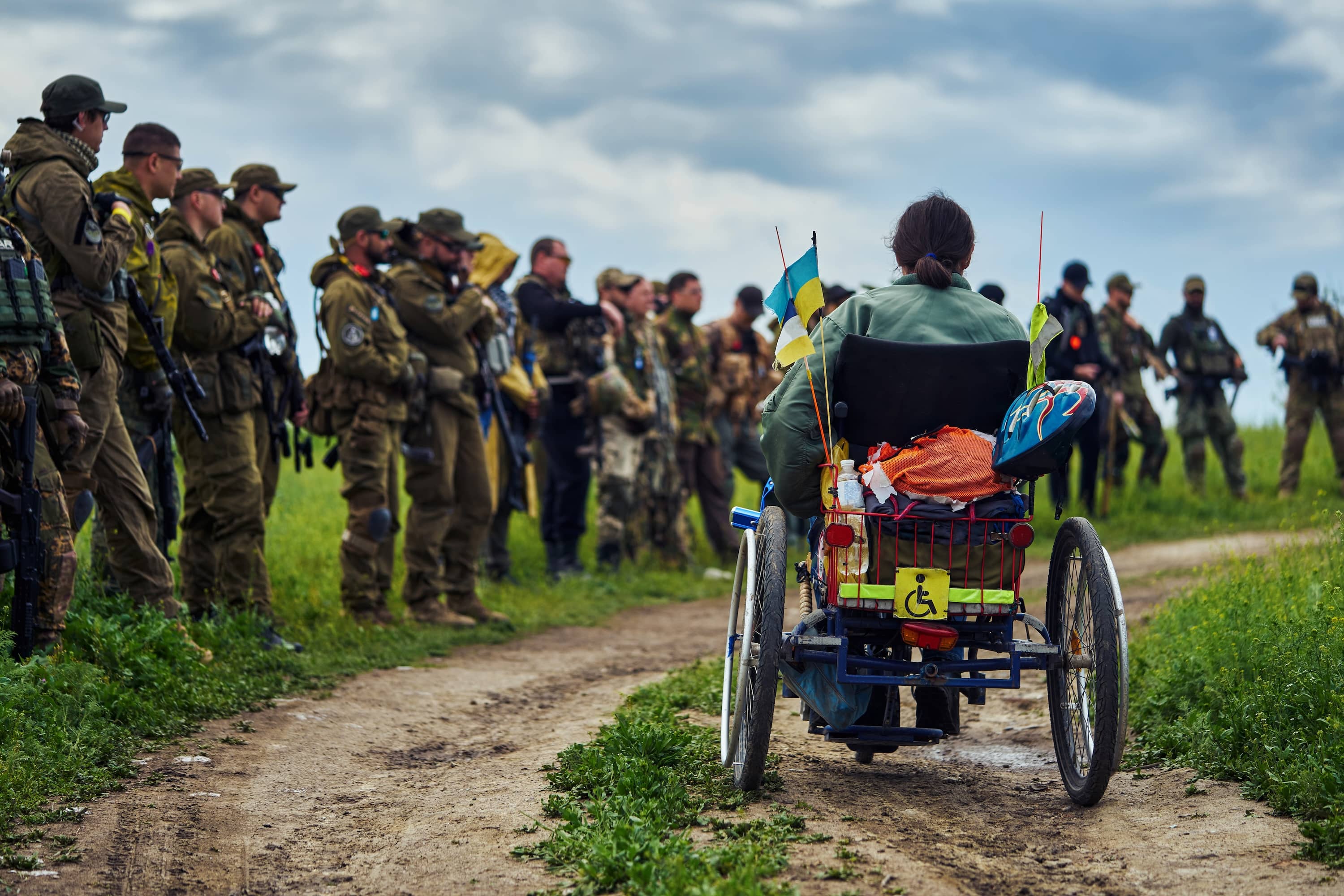 A woman in a wheelchair, with Ukrainian flags attached to it. There are armed soldiers standing on grass under a cloudy sky. 