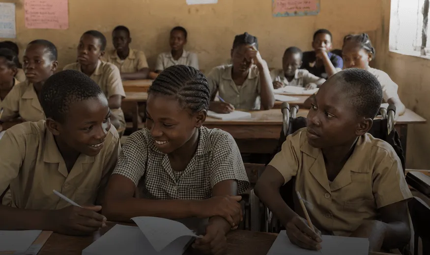 Three students smiling and writing at desks in a classroom with other students in the background. One of the children is a wheelchair user.