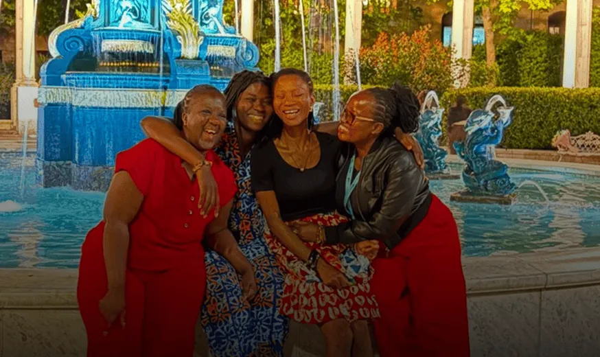 Four women smiling and hugging each other while sitting by a decorative blue fountain outdoors. One of them is a blind young girl.
