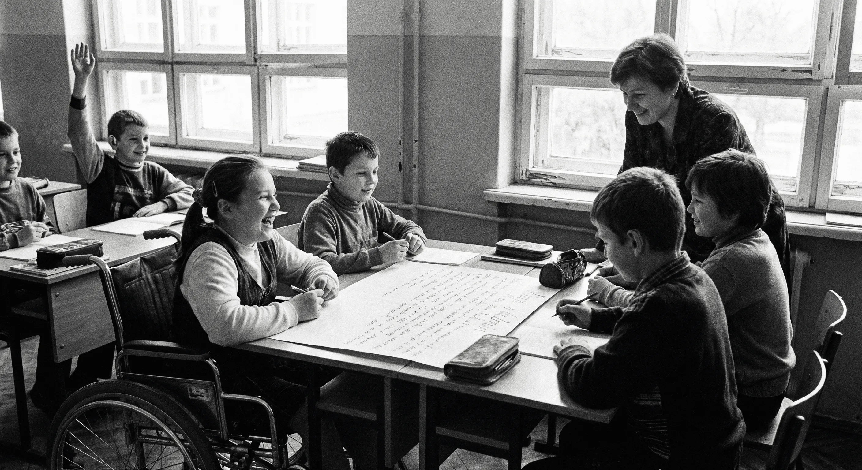 A teacher and children, including a girl in a wheelchair, smiling and working together around a table in a classroom.