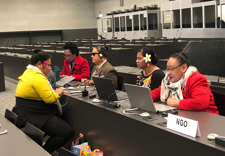 Five women seated at a long conference table with laptops and an NGO sign. There is blind woman in the group.