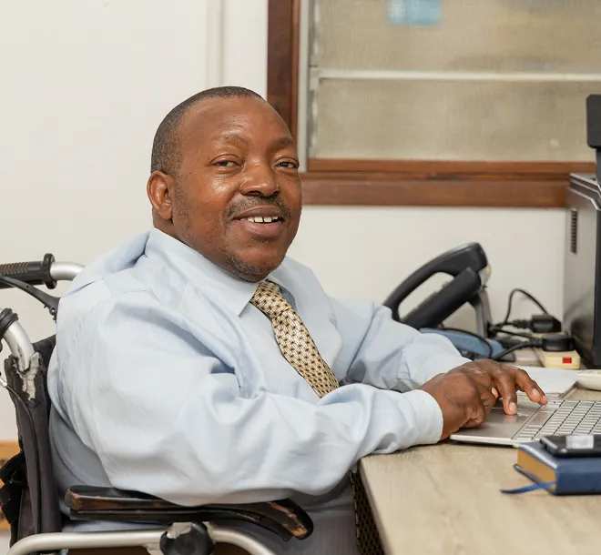 Man in a wheelchair smiling and typing on a laptop at an office desk.
