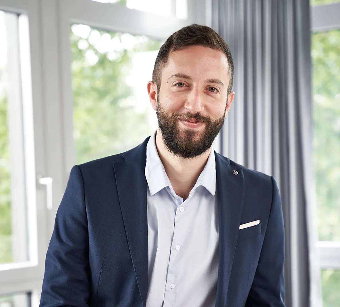Ein lächelnder Mann mit dunklem Bart trägt ein hellblaues Hemd und ein dunkelblaues Sakko vor einem Fenster mit grüner, verschwommener Landschaft.