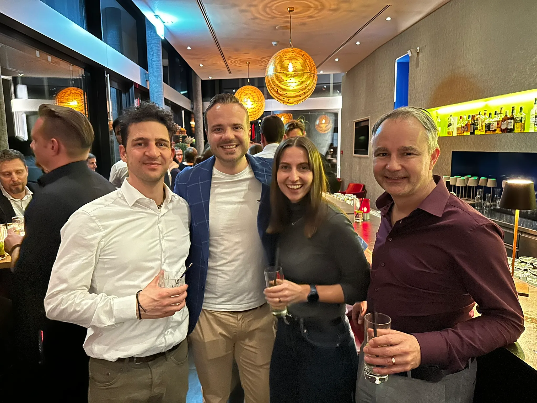 Vier Personen lächeln und halten Getränke in einer lebhaften Bar mit runden Deckenlampen im Hintergrund.