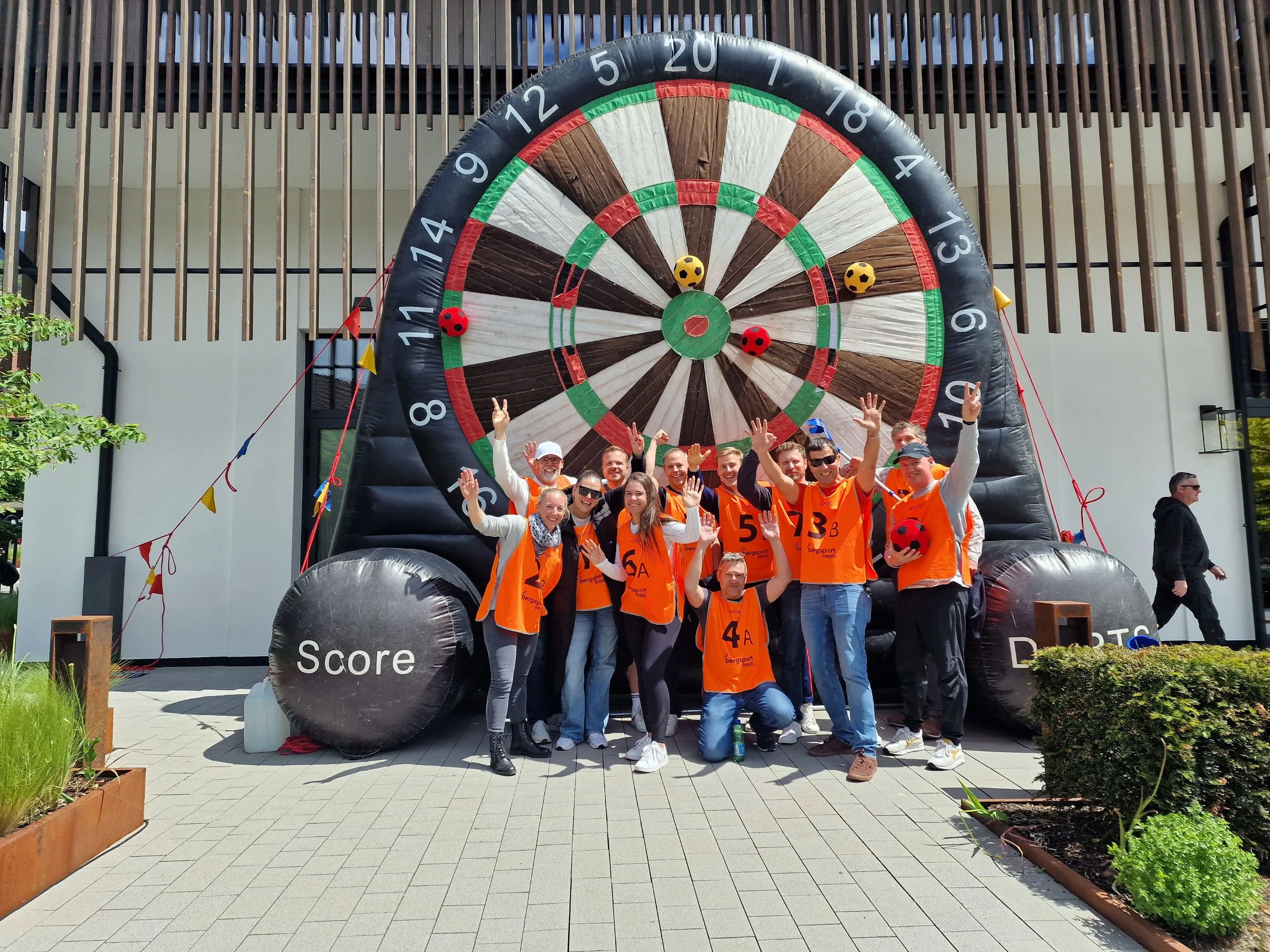 Gruppe von Menschen in orangefarbenen Trikots posiert vor einer übergroßen aufblasbaren Dartscheibe im Freien.
