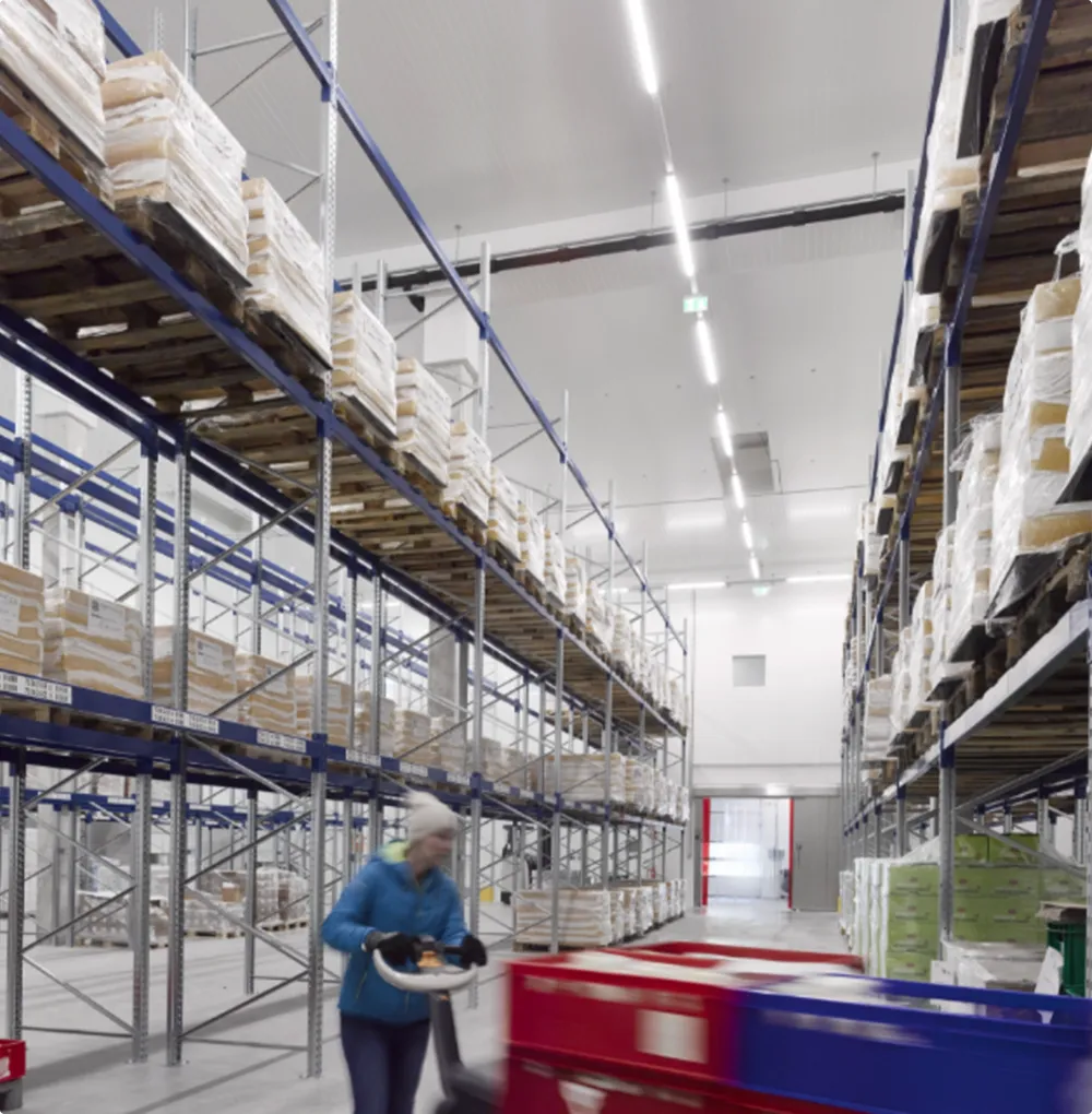 Person in einer blauen Jacke und weißer Mütze, die in einem Lagerhaus mit hohen Regalen und verpackten Paletten Kisten transportiert.