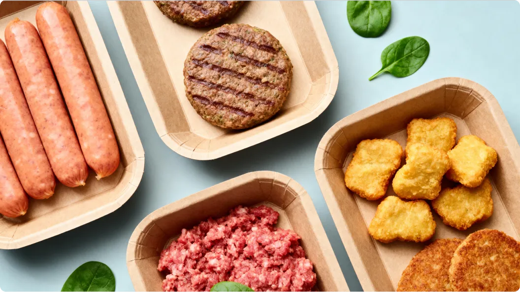 Verschiedene rohe und gekochte Fleischprodukte wie Würstchen, Burgerpattys, Nuggets und Hackfleisch in Papierschalen auf blauem Hintergrund.