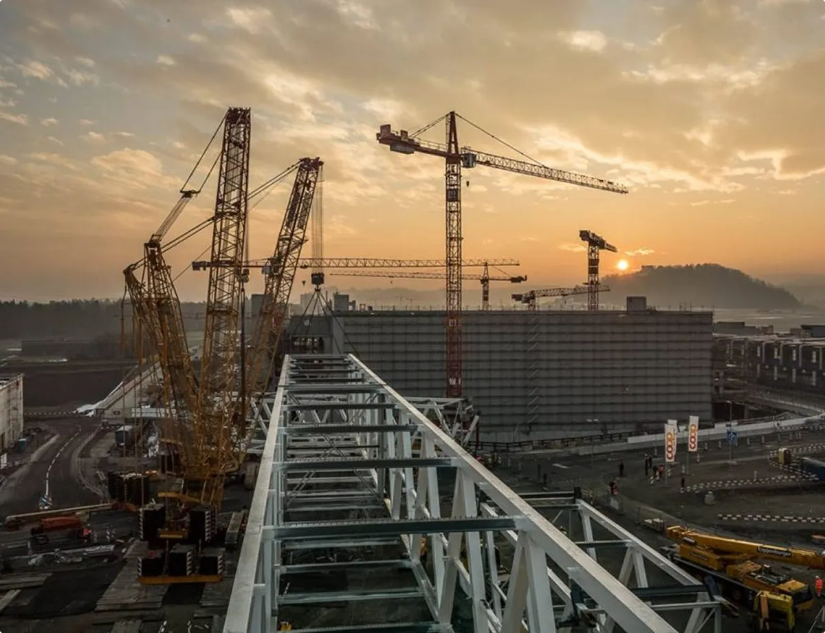 Construction site at sunset with several cranes and a large steel frame in the foreground.