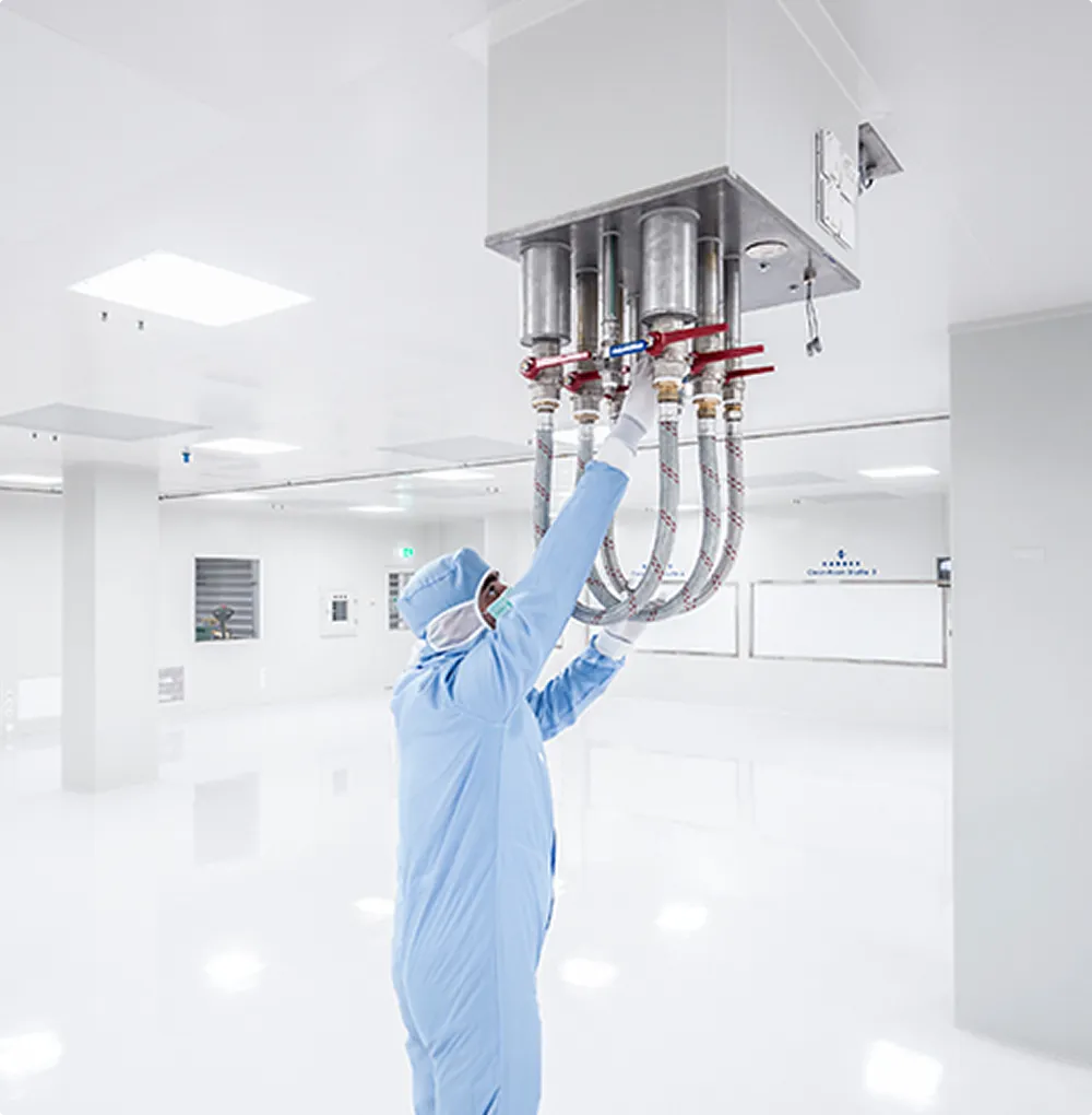 An employee wearing protective clothing checks pipes on a ceiling system in a sterile, white clean room.