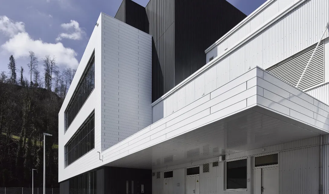 Modern, multi-storey industrial building with white and black facades in front of a wooded hill under a blue sky with clouds.