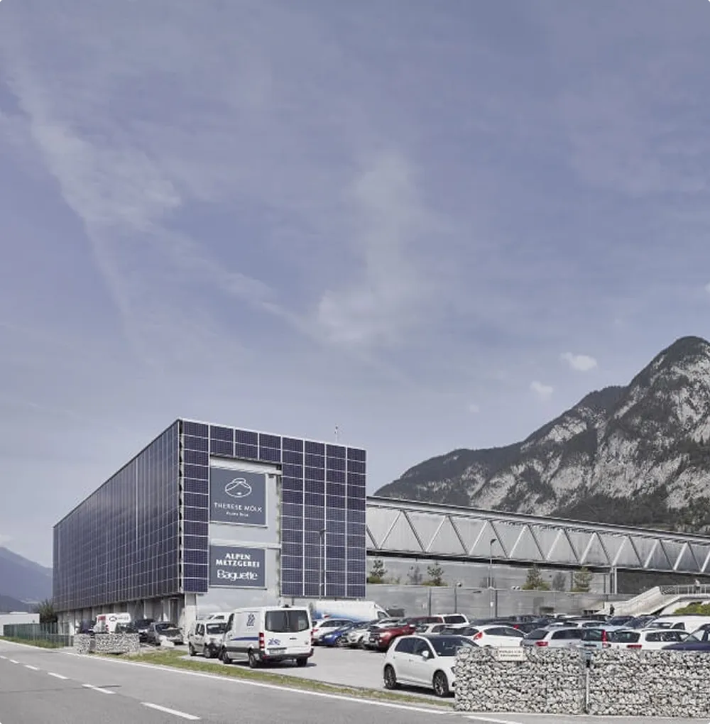 Modern industrial building with solar panels on the façade, lots of parked cars and mountains in the background.