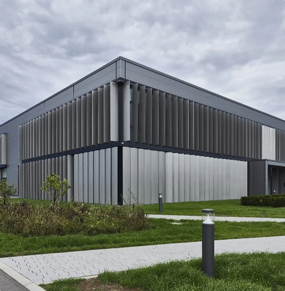 Modern grey building with vertical slats on the façade and a well-kept green area when the sky is overcast.