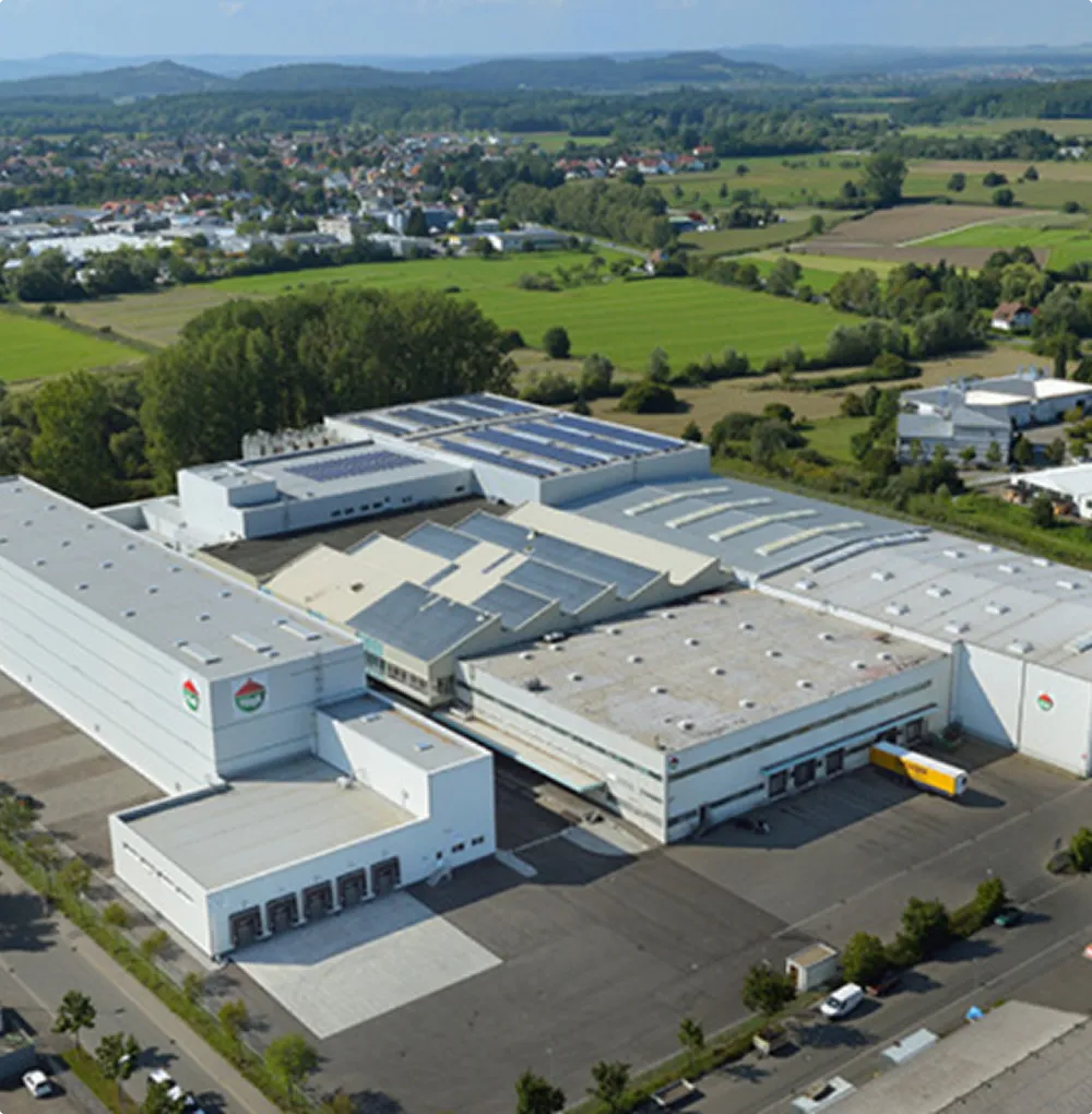 Aerial view of a large industrial building with solar panels on the roof, surrounded by green fields and rural landscape.