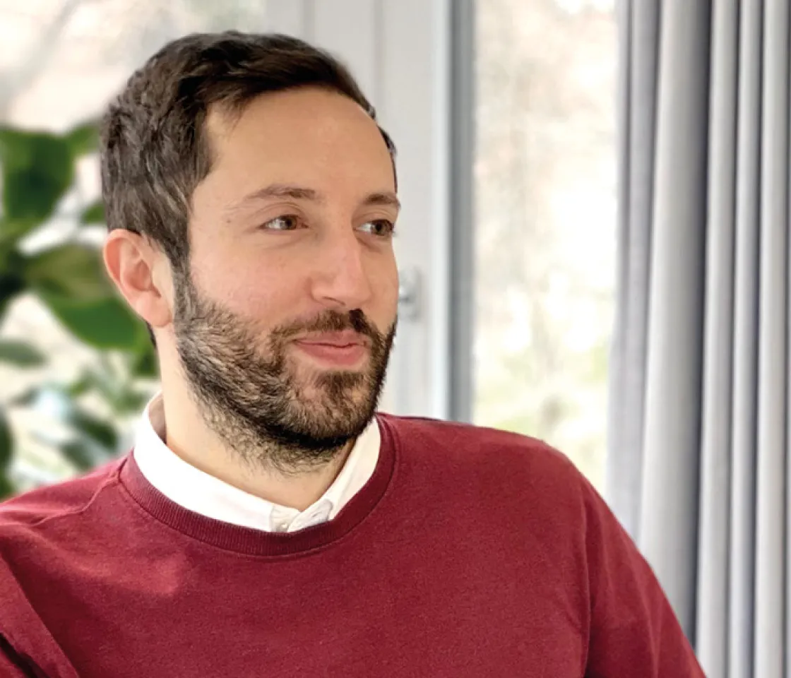 Man with short dark hair and beard wears a red sweater over a white shirt and looks to the right.
