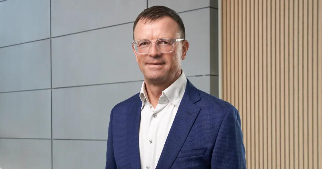 A man with short brown hair wears glasses, a white shirt and a dark blue jacket in front of a wall with light gray tiles and wooden panels.