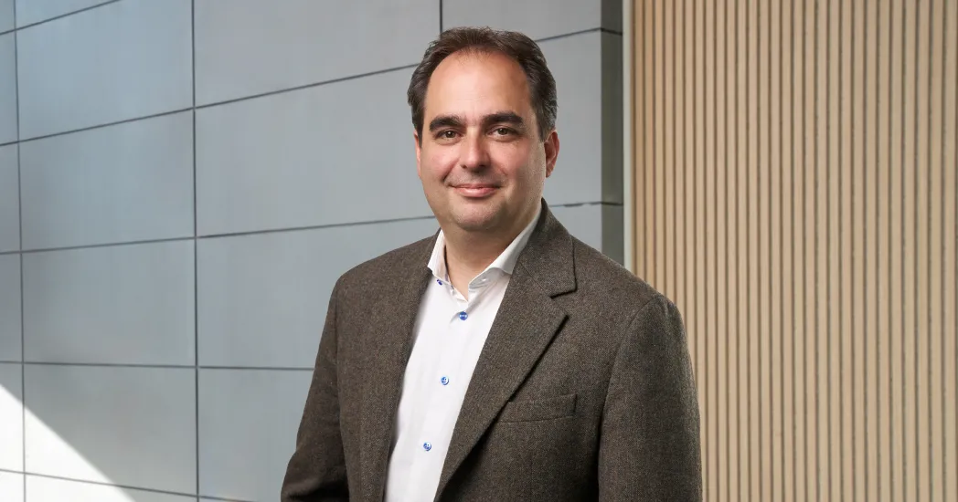 Portrait of a smiling man with dark hair in a brown blazer and white shirt against a modern wall of light panels and wood paneling.