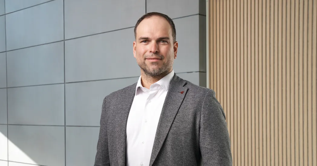 A man in a gray jacket and white shirt stands in front of a wall with gray tiles and vertical wooden boards.