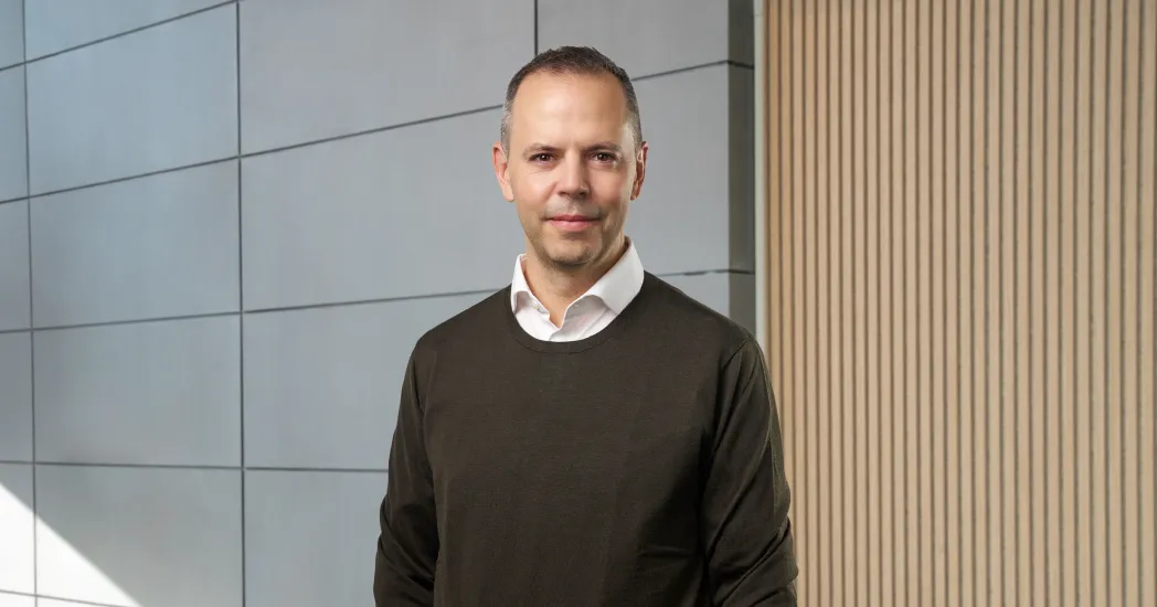 Man wearing a brown sweater and white shirt in front of a grey and beige wall.