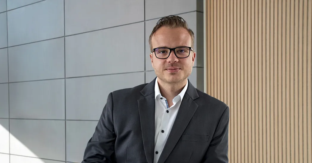 Portrait of a man wearing glasses in a black suit and white shirt in front of a wall with tiles and wood paneling.