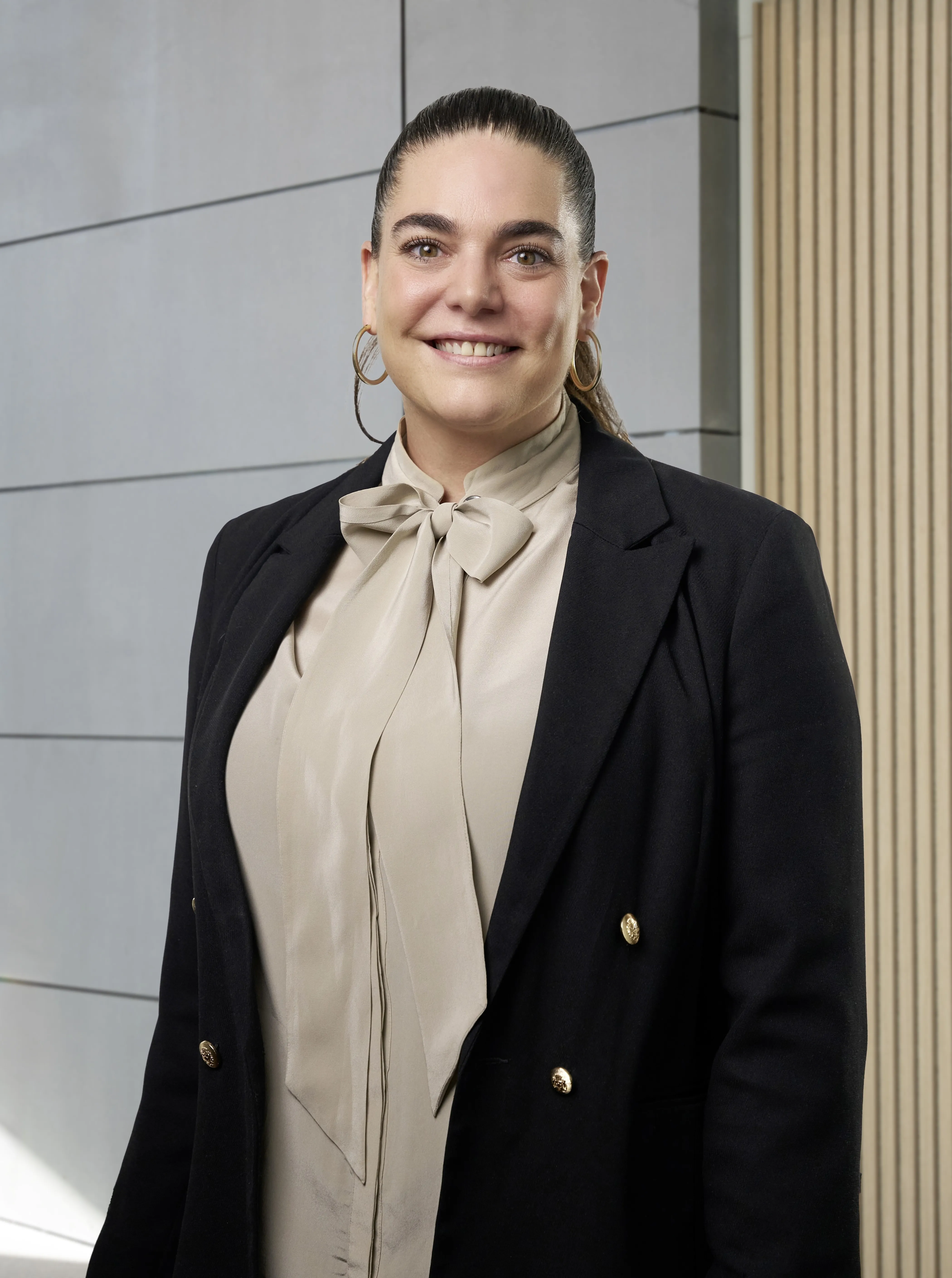 Frau mit braunen Haaren im Pferdeschwanz trägt eine beige Bluse mit Schleife und einen schwarzen Blazer mit goldenen Knöpfen, lächelt vor einem modernen Gebäude.