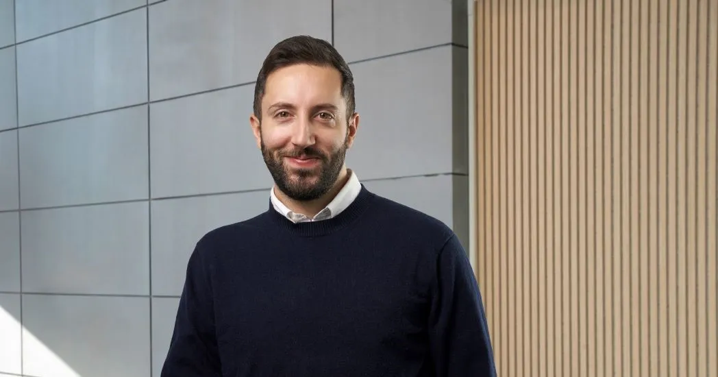 A smiling man with a dark sweater and beard stands in front of a wall with gray tiles and a wooden panel wall.