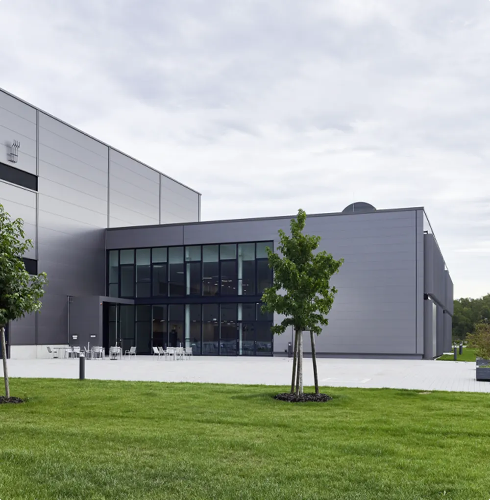 Modern grey industrial building with large windows, in front of it a paved area with tables and chairs and a green lawn with young trees.