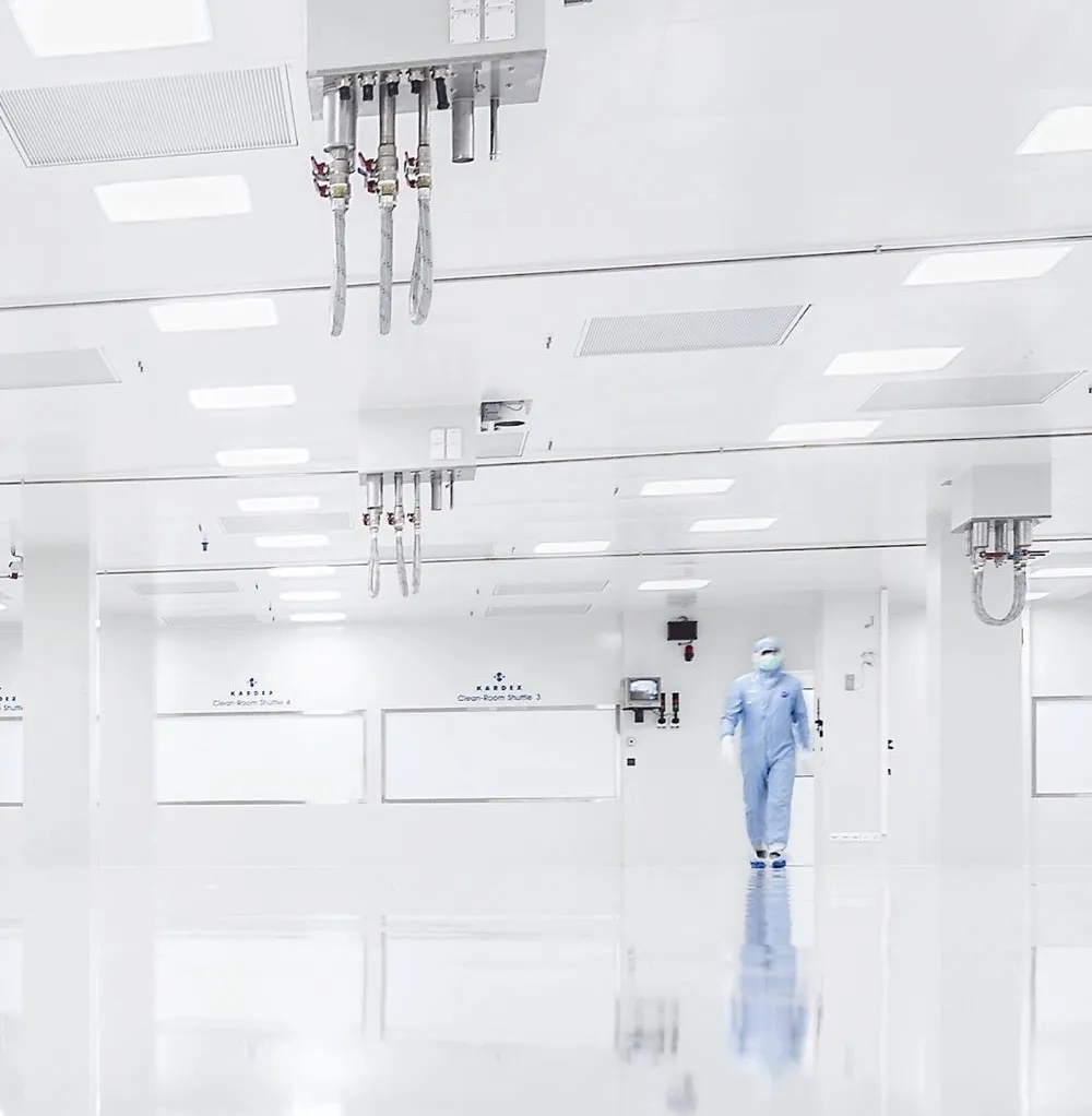 Person in Schutzanzug und Maske geht durch einen sterilen, weißen Reinraum mit reflektierendem Boden.