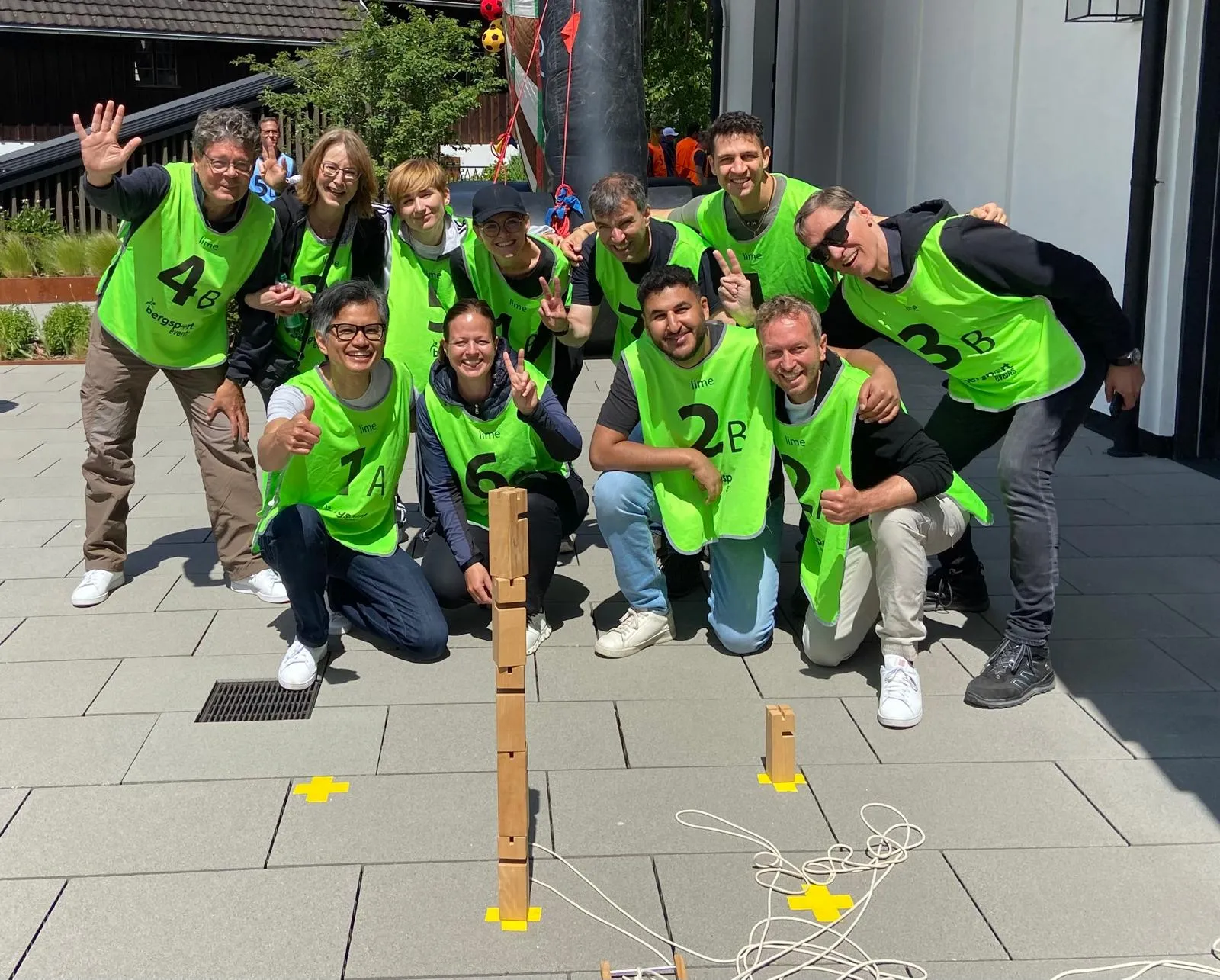 Elf Personen in grünen Sportwesten posieren lächelnd im Freien hinter einem Turm aus Holzblöcken auf dem Boden.