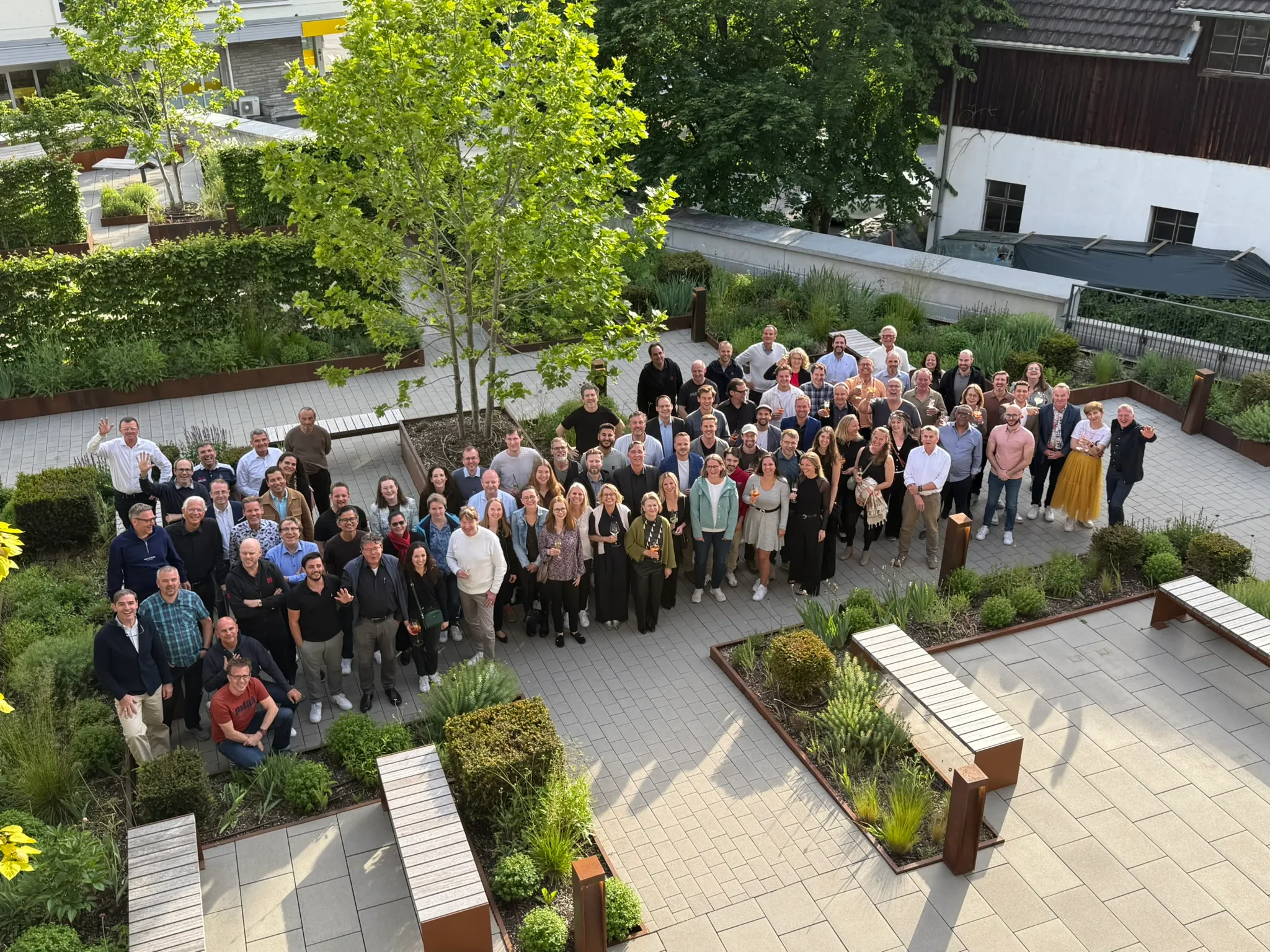 Große Gruppe von Menschen versammelt in einem begrünten Hof mit Sitzbänken, die in die Kamera lächeln und winken.
