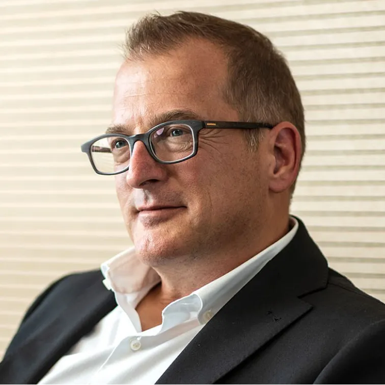 Porträt eines lächelnden Mannes mit Brille, weißem Hemd und dunklem Anzug vor einer hellen, gestreiften Wand.