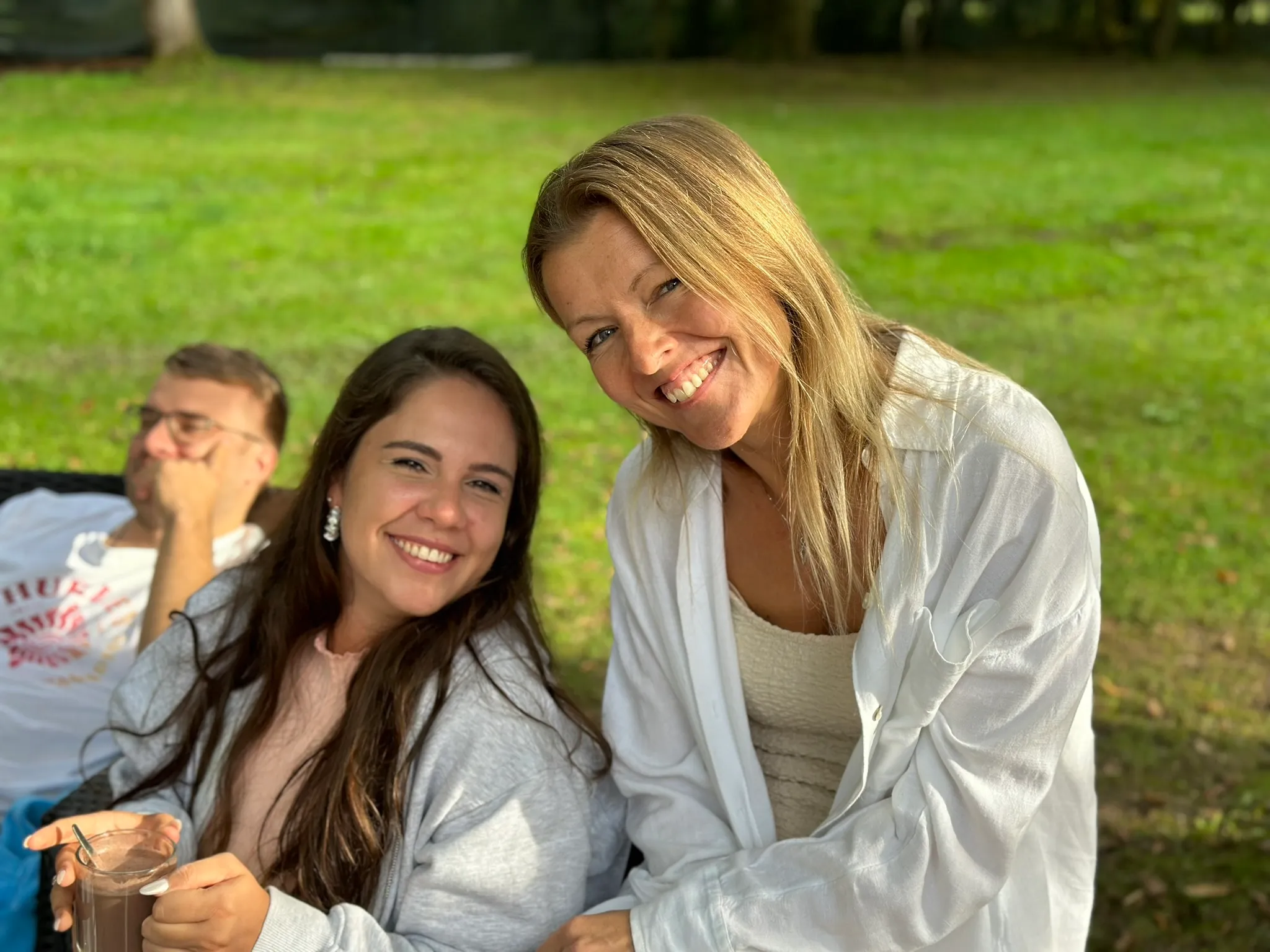 Zwei lächelnde Frauen, eine mit dunklen langen Haaren hält ein Glas mit Schokoladenmilch, im Hintergrund sitzt ein Mann auf einer grünen Wiese.
