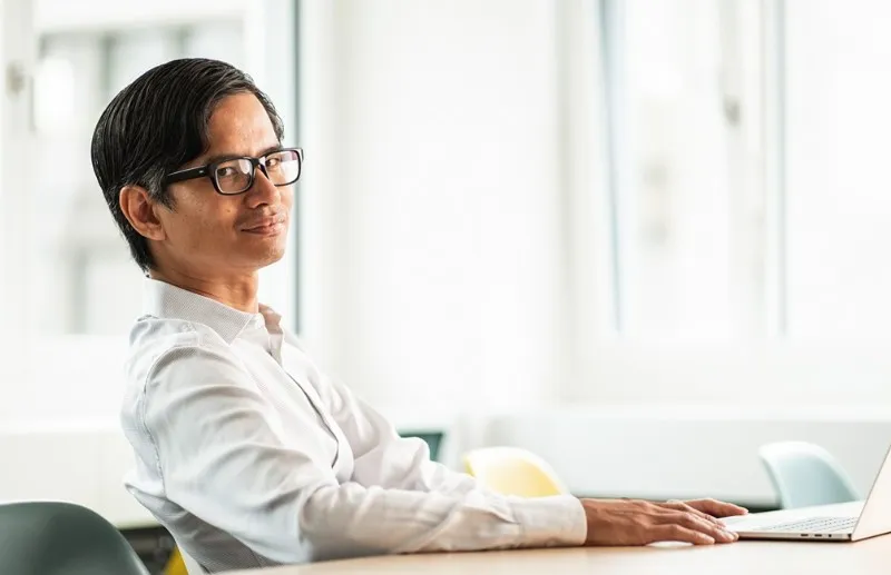 Mann mit schwarzer Brille sitzt entspannt an einem Tisch mit Laptop in einem hellen Raum.