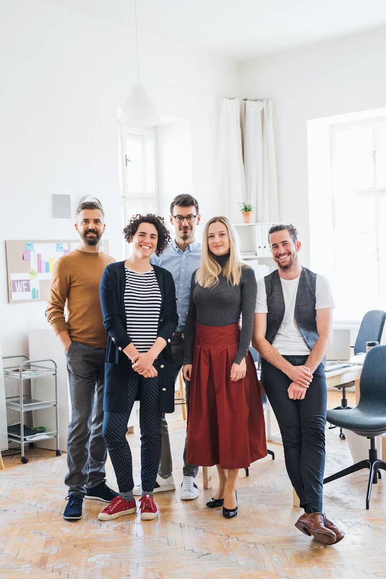 Five casually dressed young professionals standing and smiling in a bright office space with wooden floors and large windows.