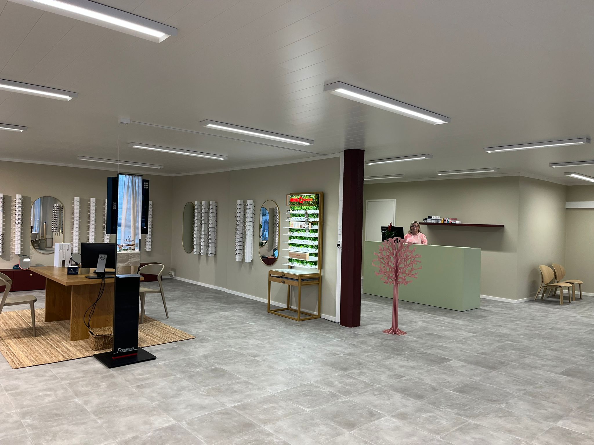 Interior of an optician's store with eyeglass displays on walls, a wooden table with chairs, and a receptionist standing behind a counter.