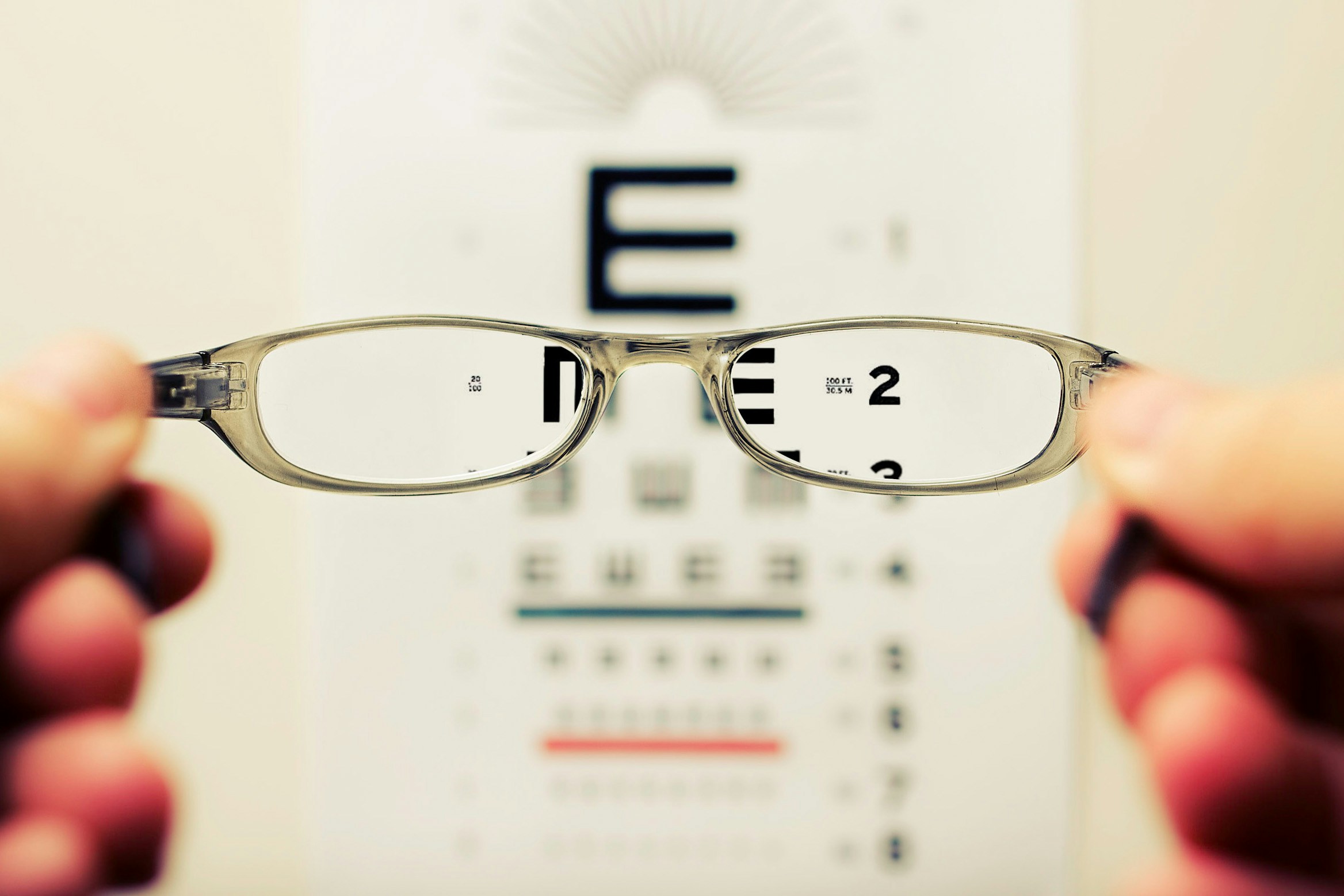 Clear eyeglasses held in front of an eye chart, showing sharp focus through the lenses while the chart is blurred around them.