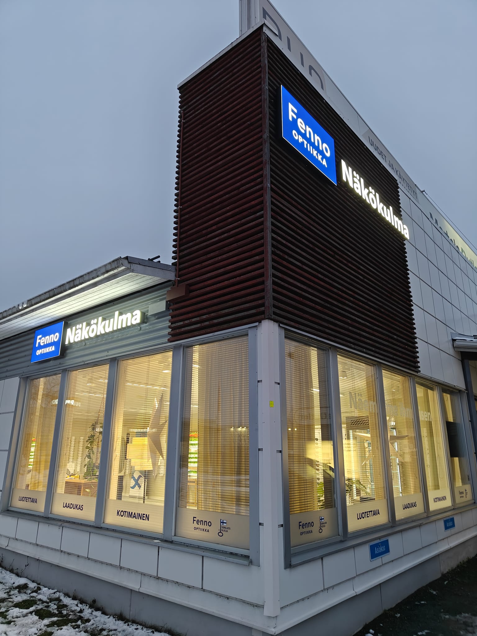 Corner view of a Fenno Optiikka Näkökulma eyewear store with illuminated signs and large windows showing the interior lighting.