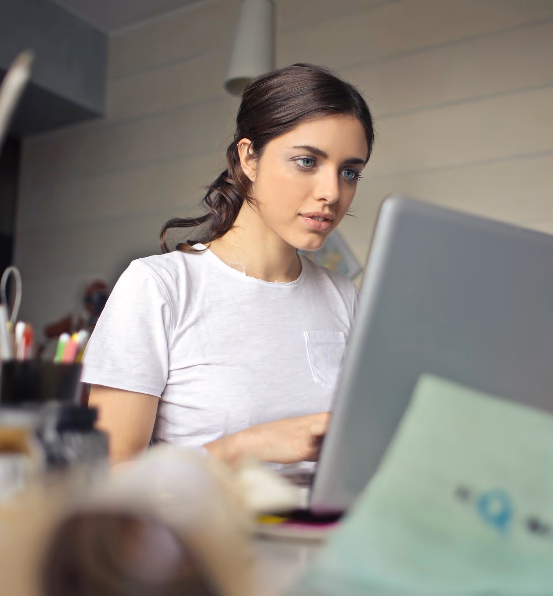 Young woman with dark hair tied back wearing a white t-shirt working on a laptop in a room.