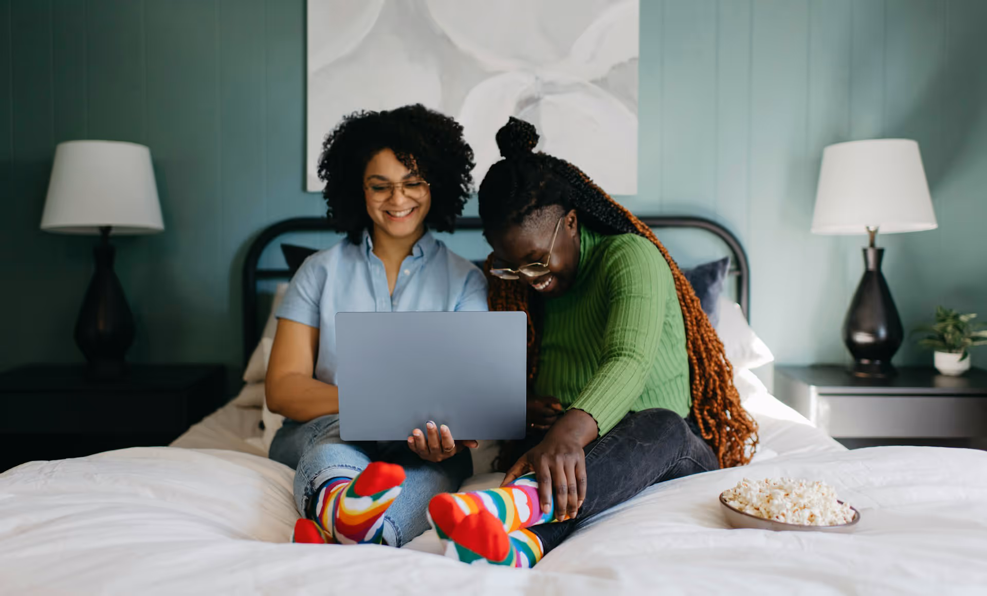 Two women sitting on a bed smiling and looking at a laptop, wearing colorful rainbow socks, with a bowl of popcorn nearby.