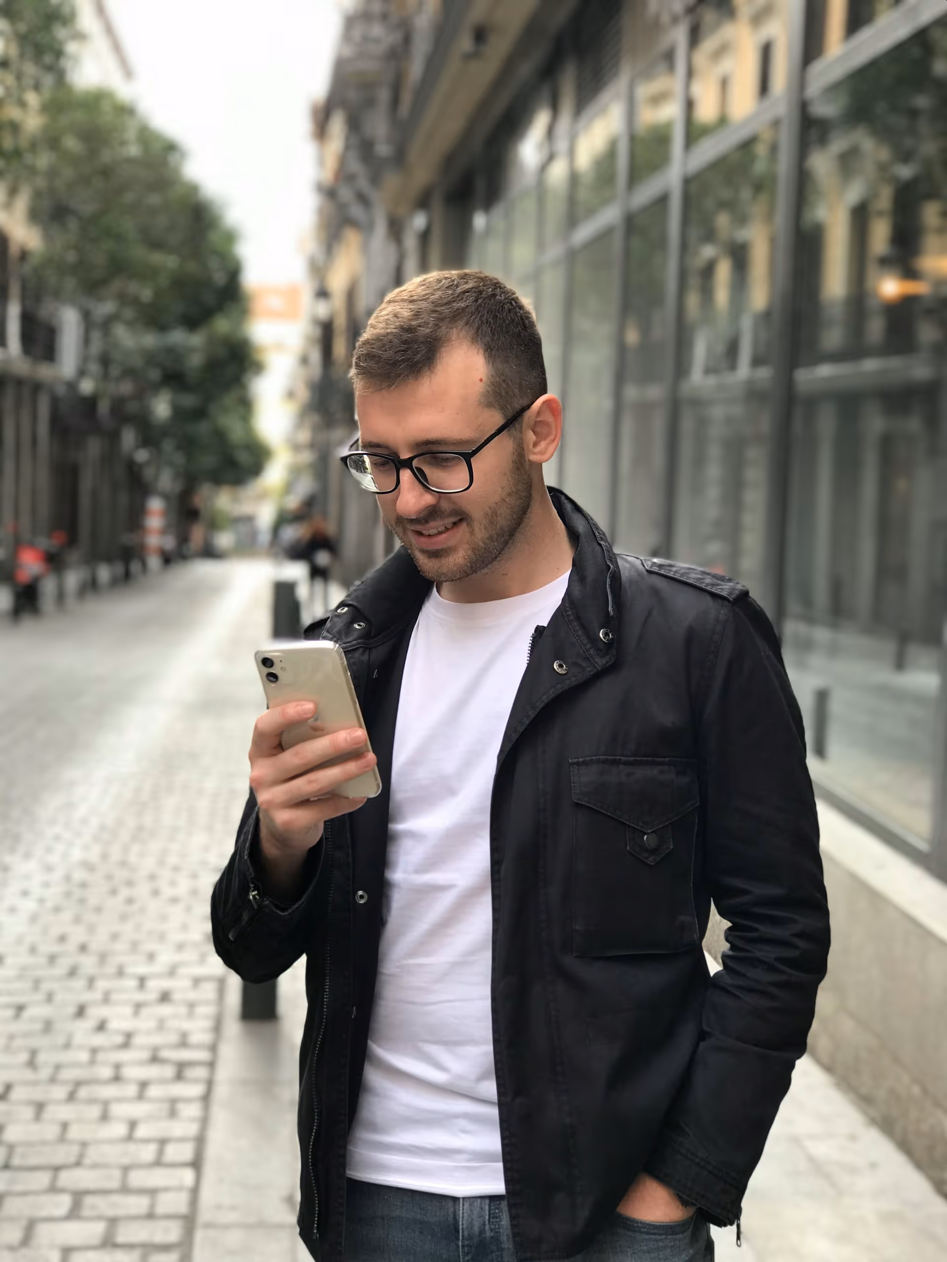 Smiling man wearing glasses, a black jacket, and white t-shirt looking at his smartphone on a city street.