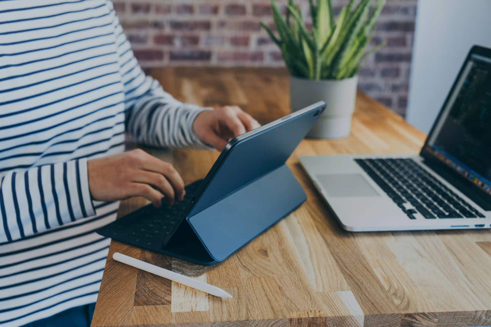 Person typing on a tablet with keyboard cover at a wooden desk, next to a laptop and white stylus pen.