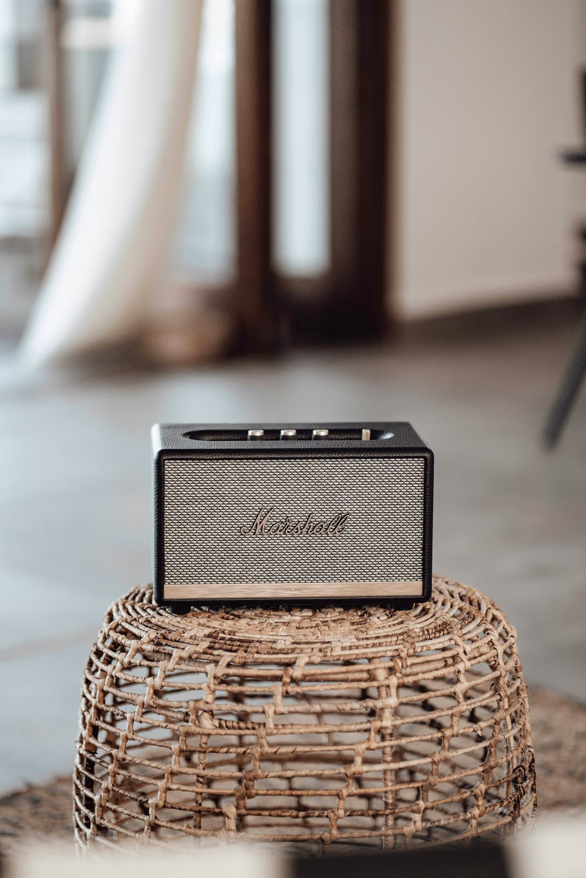 Black and gold Marshall speaker resting on a woven wicker stool in a softly lit room.